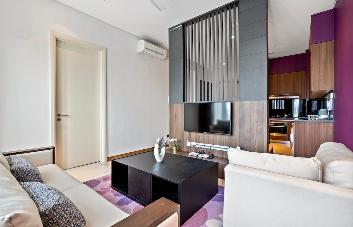 The living area is designed with a stylish touch, featuring a modern entertainment unit with a large flat-screen TV. Soft furnishings include two sofas, a coffee table with decorative items, and a backdrop of rich purple walls that contrast with neutral tones.