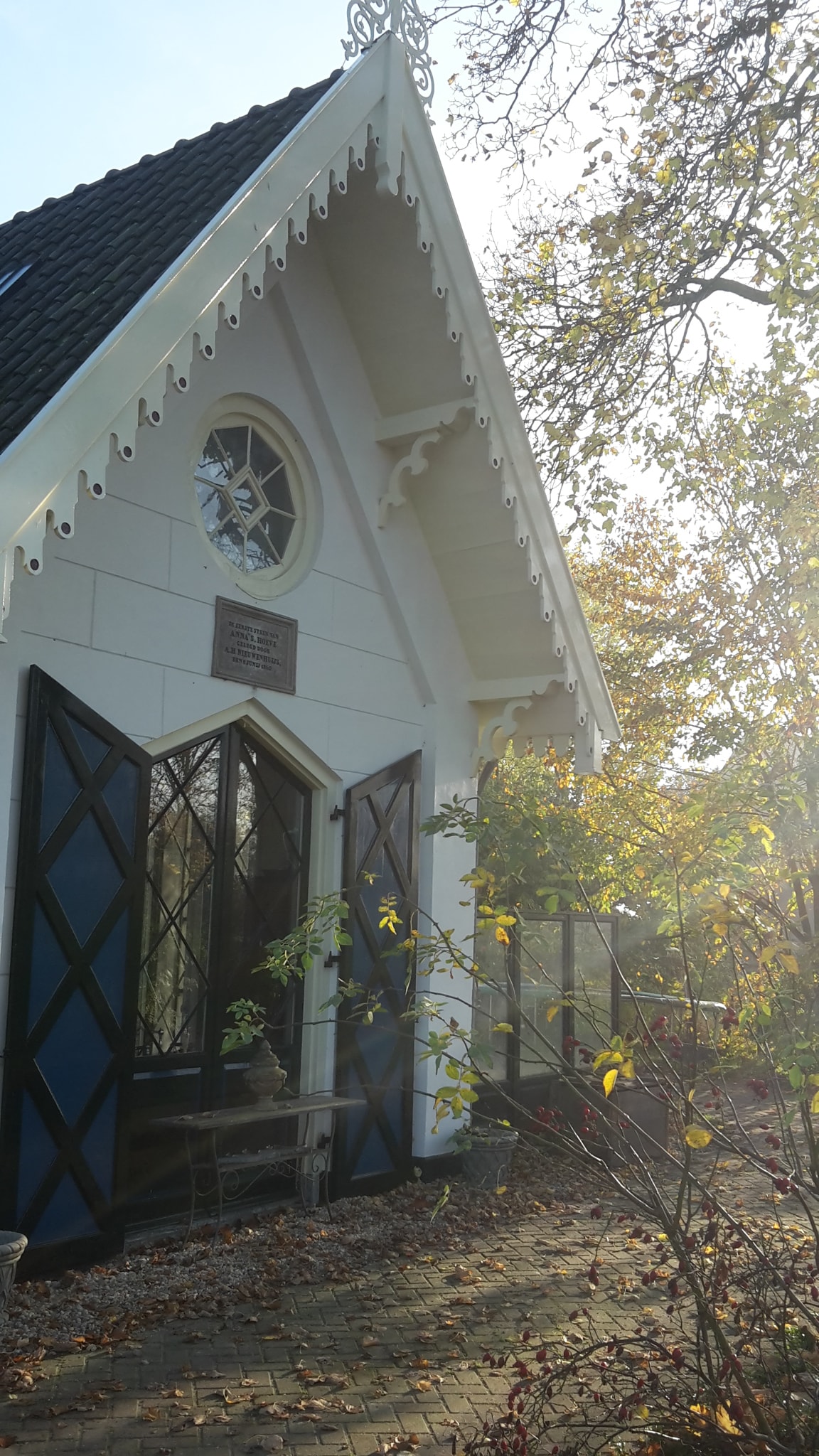 A charming exterior of a house is framed by trees, showcasing decorative gables and large windows with blue shutters. The light filters through the leaves, creating a warm, inviting ambiance around the entrance.