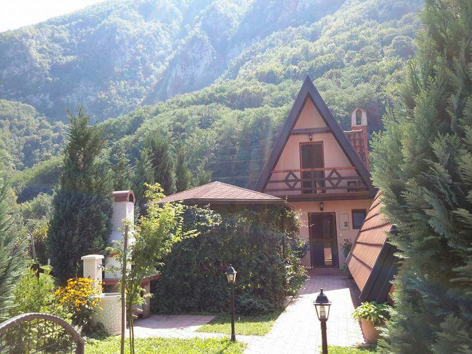 A charming bungalow with an A-frame roof is surrounded by tall trees and vibrant greenery. A paved pathway leads to the entrance of the home, which features a cozy porch and a small garden with seasonal flowers. Mountain peaks rise majestically in the background.
