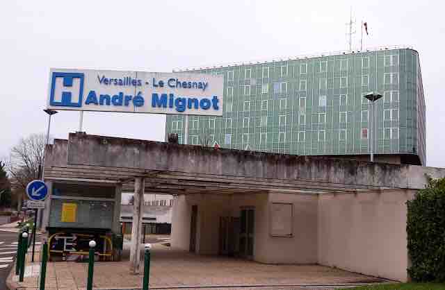 The image displays the entrance of the André Mignot Hospital, featuring a large, illuminated sign with the hospital's name. A modern building with a green glass façade is visible in the background. The surrounding area includes a parking space and street signage.