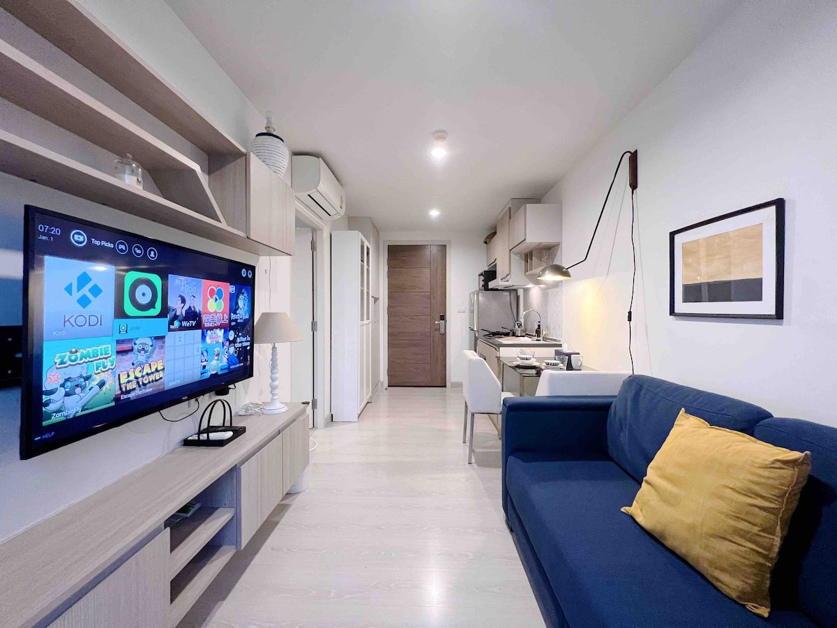 A modern living area features a navy blue sofa with a yellow accent pillow positioned in front of a wall-mounted TV. Shelving units display decorative items, while a small dining area is seen in the background, accompanied by a well-equipped kitchen.