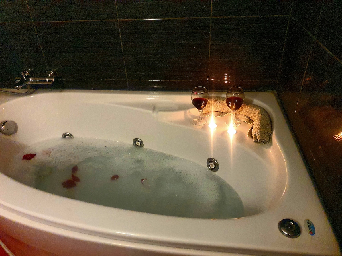 The bathroom features a modern jacuzzi bath, filled with bubbles and adorned with rose petals. Two glasses of wine rest nearby, accompanied by a lit candle, contributing to a serene atmosphere.