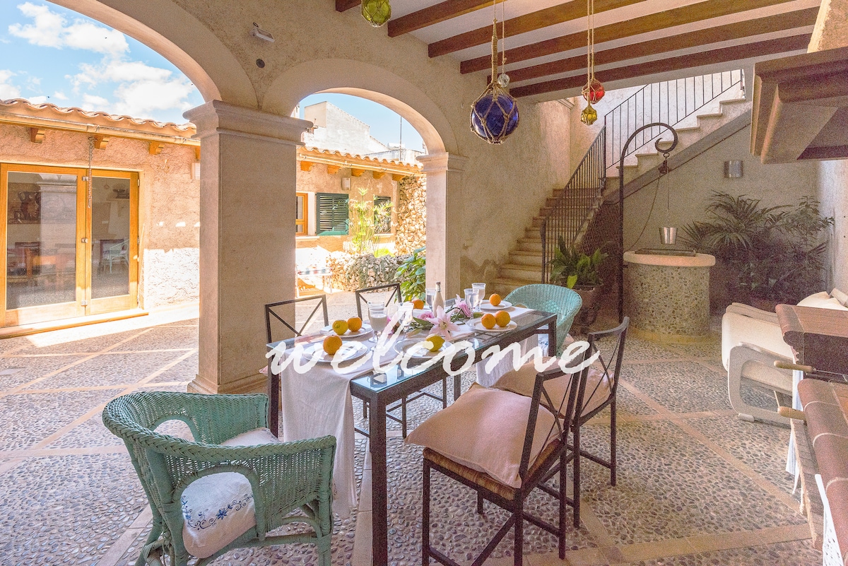 An outdoor dining area is depicted, featuring a table set with fruit and soft seating. Surrounding this space are stone arches and decorative accents, while the staircase in the background leads to an upper level. Natural light illuminates the scene, enhancing the inviting atmosphere.