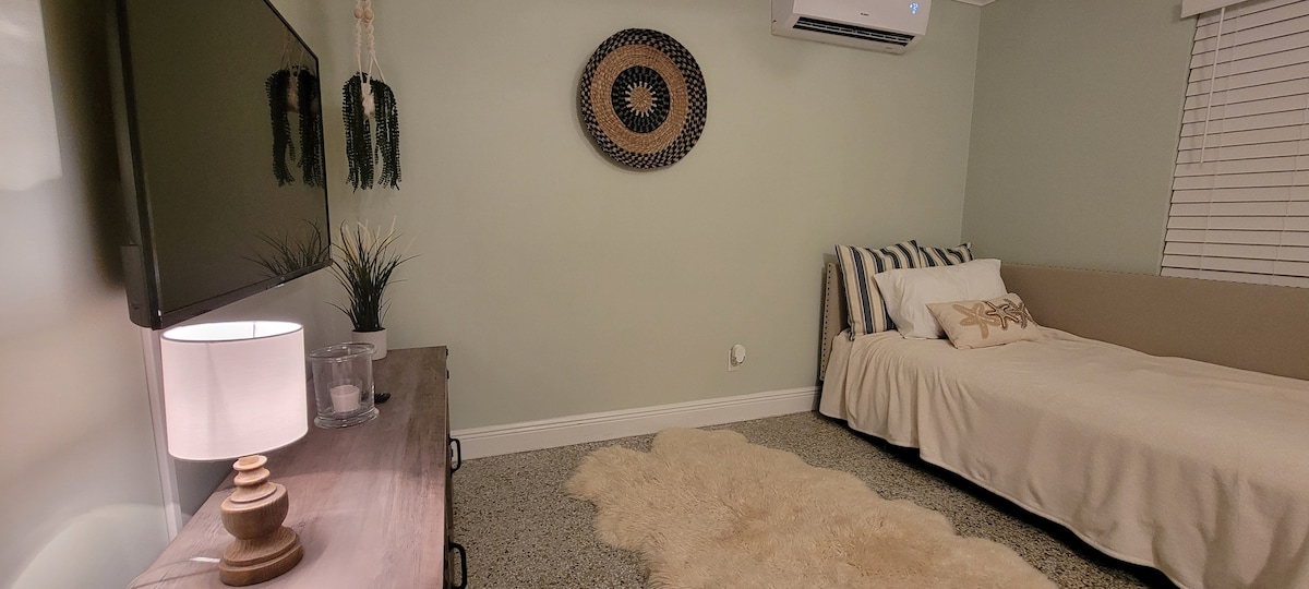 A calm living space features a twin daybed adorned with neutral-colored bedding, alongside a light wooden dresser with a lamp. A textured area rug lays on the floor, while wall decor includes a circular woven piece. An air conditioning unit is quietly positioned above.