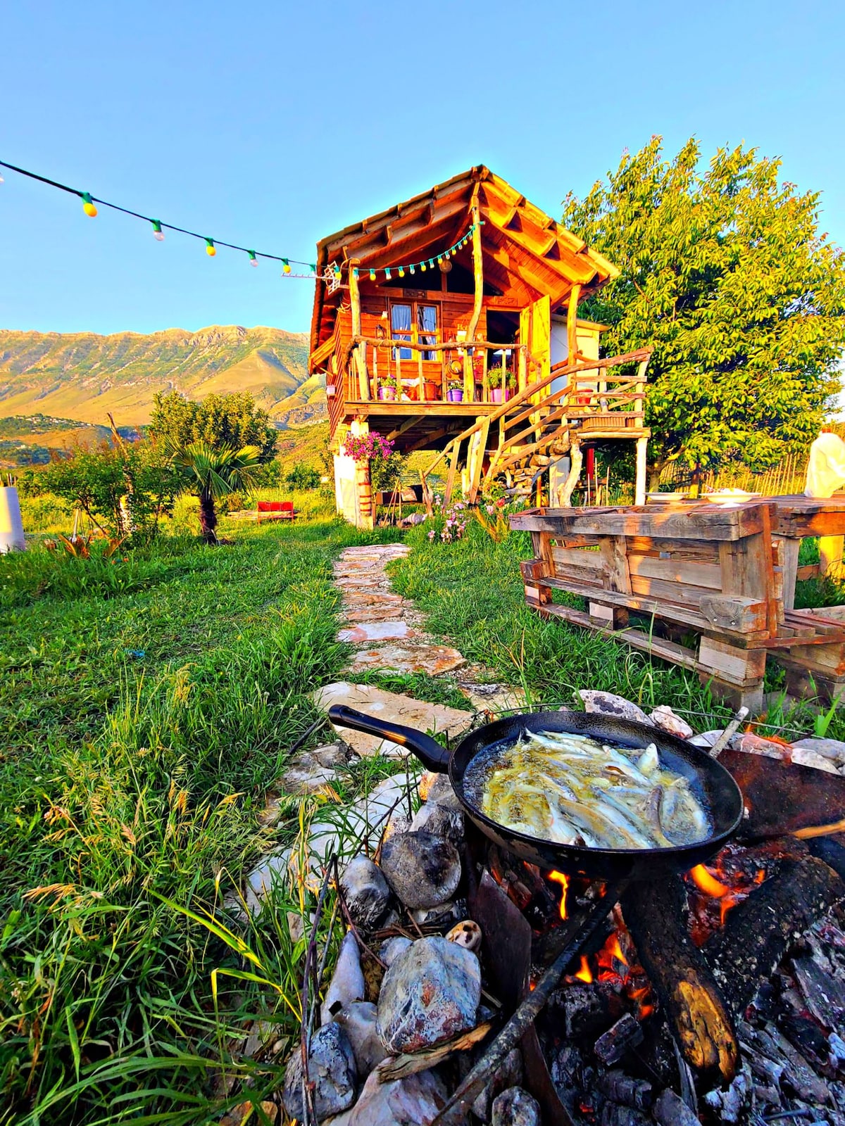 A two-story wooden structure is surrounded by natural greenery, illuminated by decorative string lights. A pathway leads to the building, while a cooking area is visible in the foreground, featuring a black pan over a fire with fish being prepared.