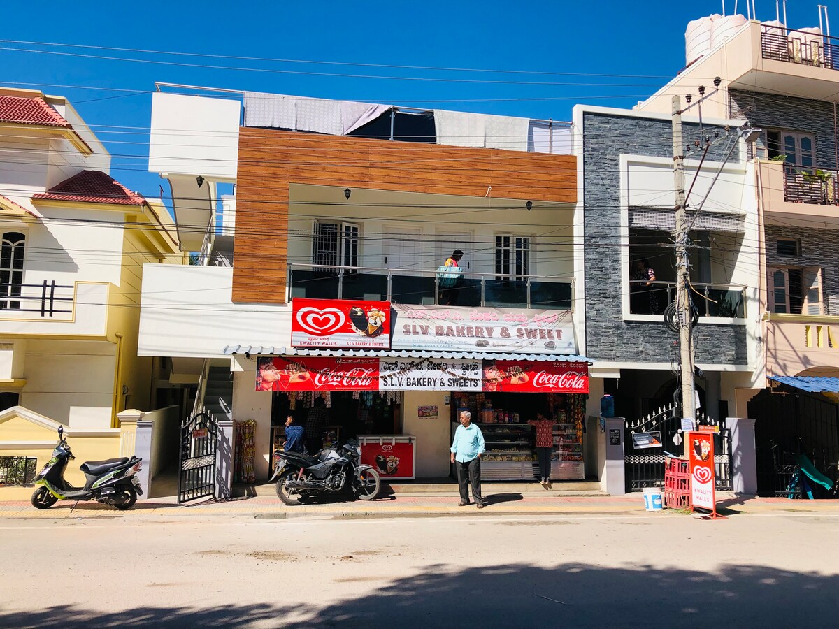 A two-story building in a vibrant neighborhood is visible, featuring a bakery and sweet shop on the ground floor. The shop's front is adorned with colorful signage and a variety of products. Motorcycles are parked nearby, and residents can be seen on the upper floor balcony.