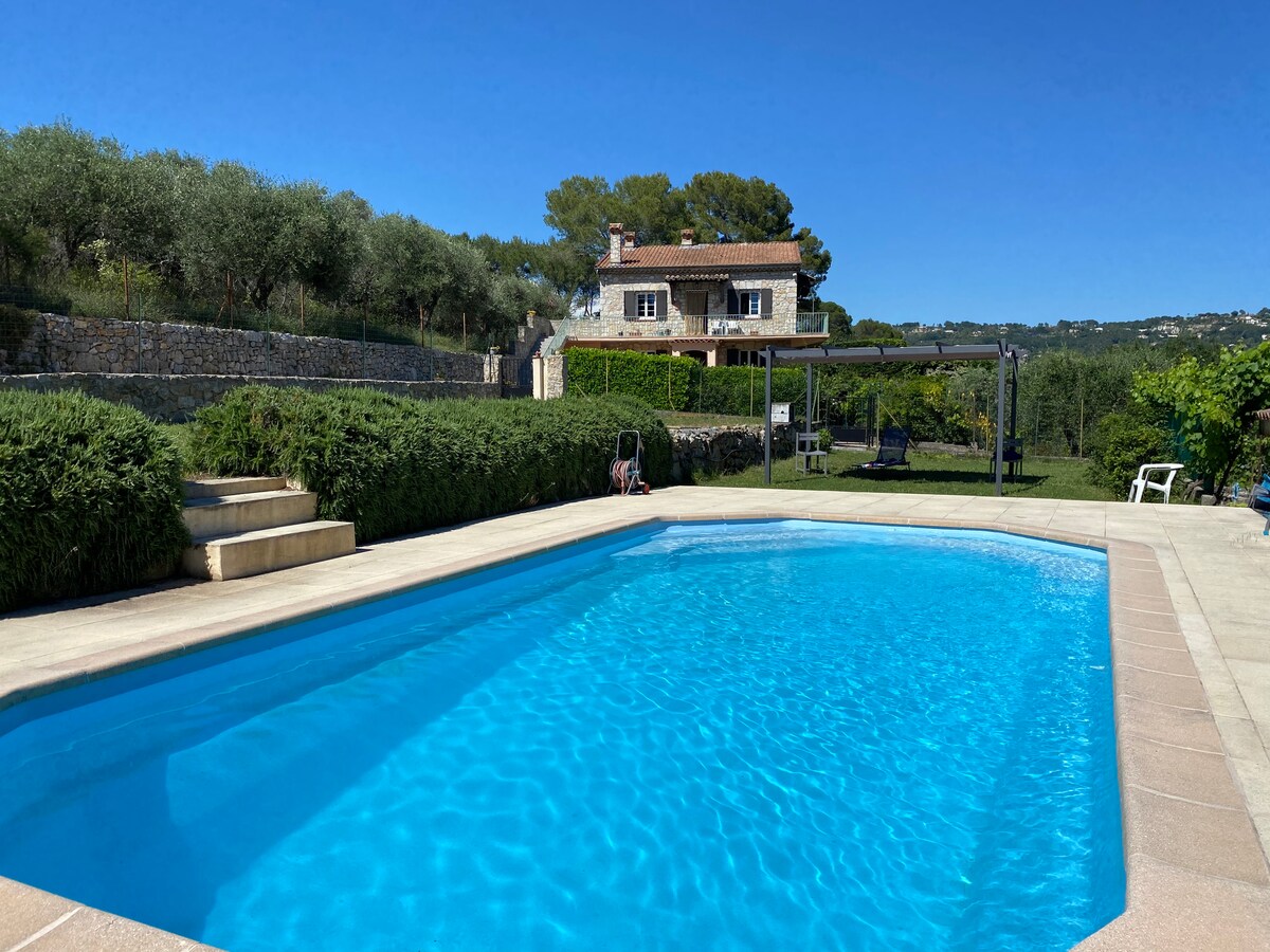 A clear blue salt-water pool is surrounded by a well-maintained garden with olive trees. Steps lead from the pool area to a grassy expanse. The villa is visible in the background, framed by greenery, under a bright blue sky.