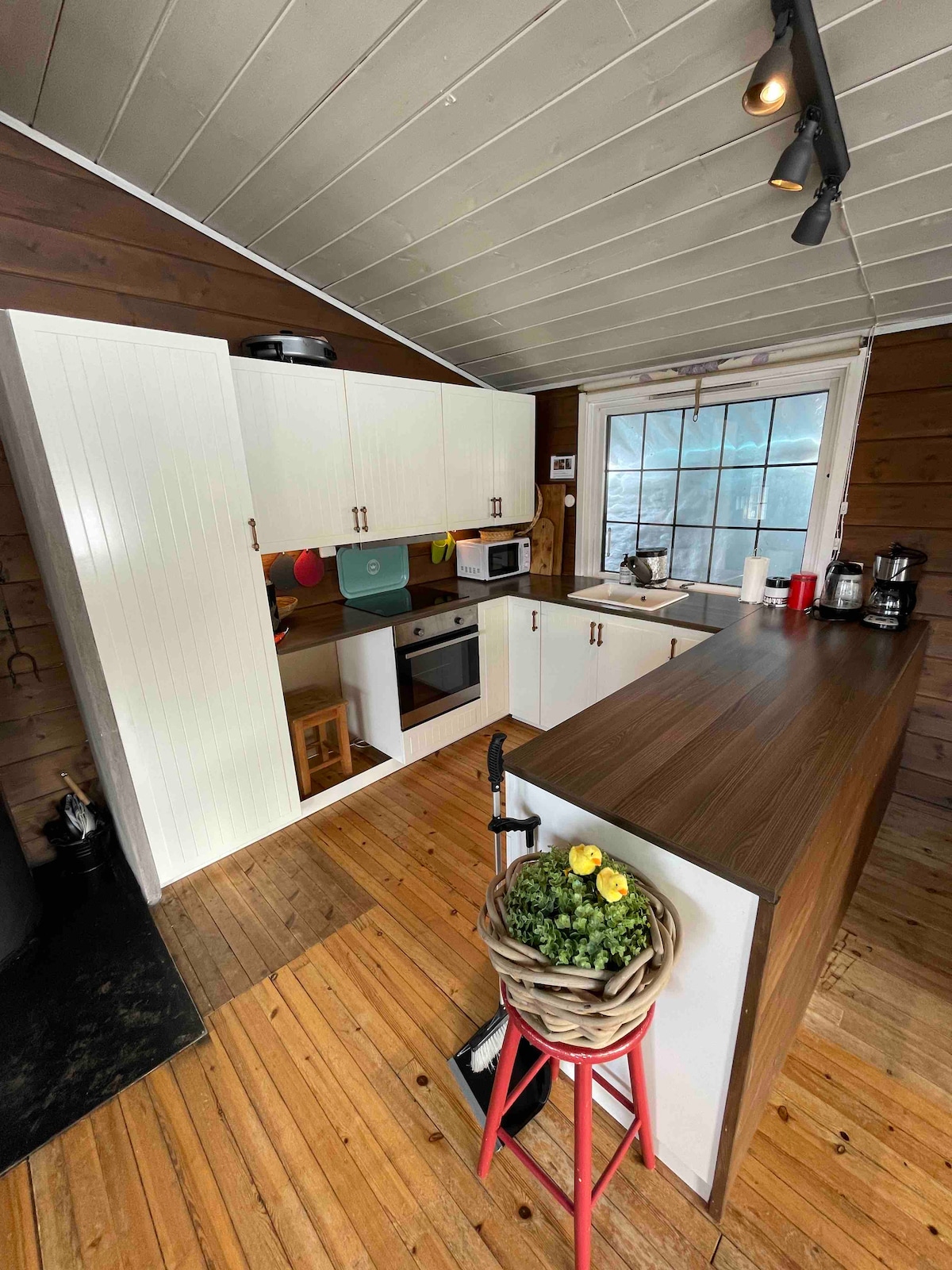 The kitchen features a wooden countertop with a sink and various appliances, including a microwave and coffee maker. White cabinets with colorful accessories provide storage. A large window allows natural light to illuminate the space, highlighting the warm wooden flooring.