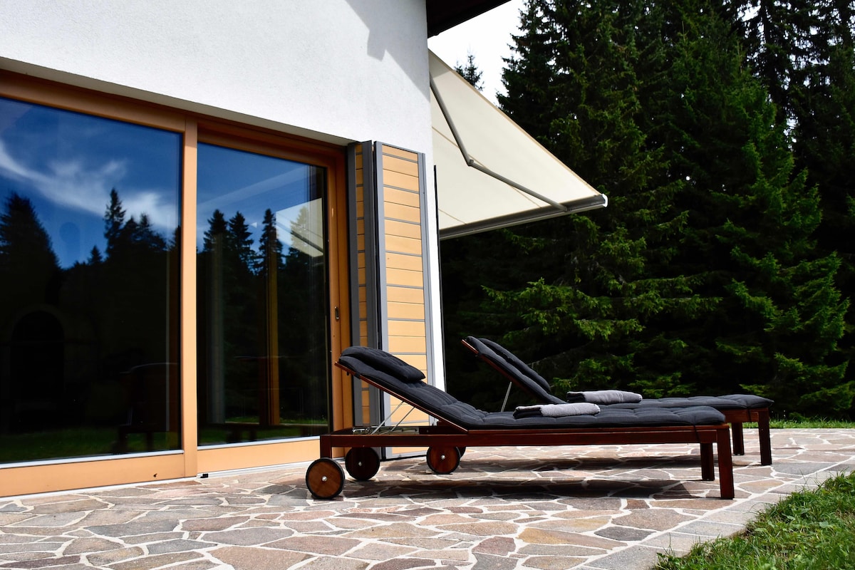 Two lounge chairs are positioned on a stone patio, offering a view of the surrounding forest. Large glass doors reflect the greenery outside, while an awning provides shade. The scene is framed by tall trees, contributing to a serene and natural atmosphere.