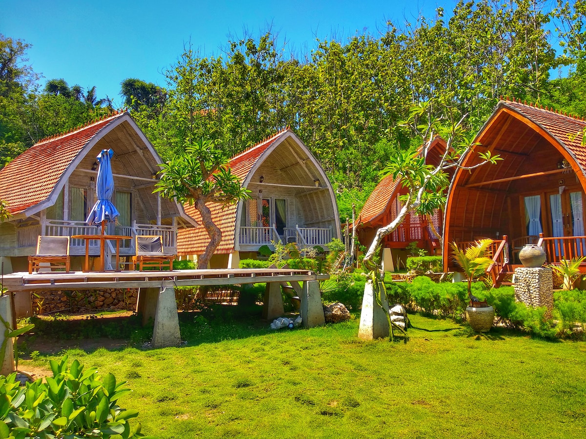 Three wooden cottages are nestled in a lush green setting, featuring sloped roofs with terracotta tiles. Each cottage has a small terrace, with outdoor seating visible. Tropical plants and trees surround the area, providing a serene environment under a clear blue sky.