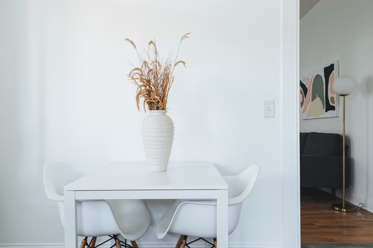A simple dining area features a white table with a light vase filled with dried grass. Two white chairs complement the table. In the background, a living area is suggested with a dark couch and abstract wall art partially visible.