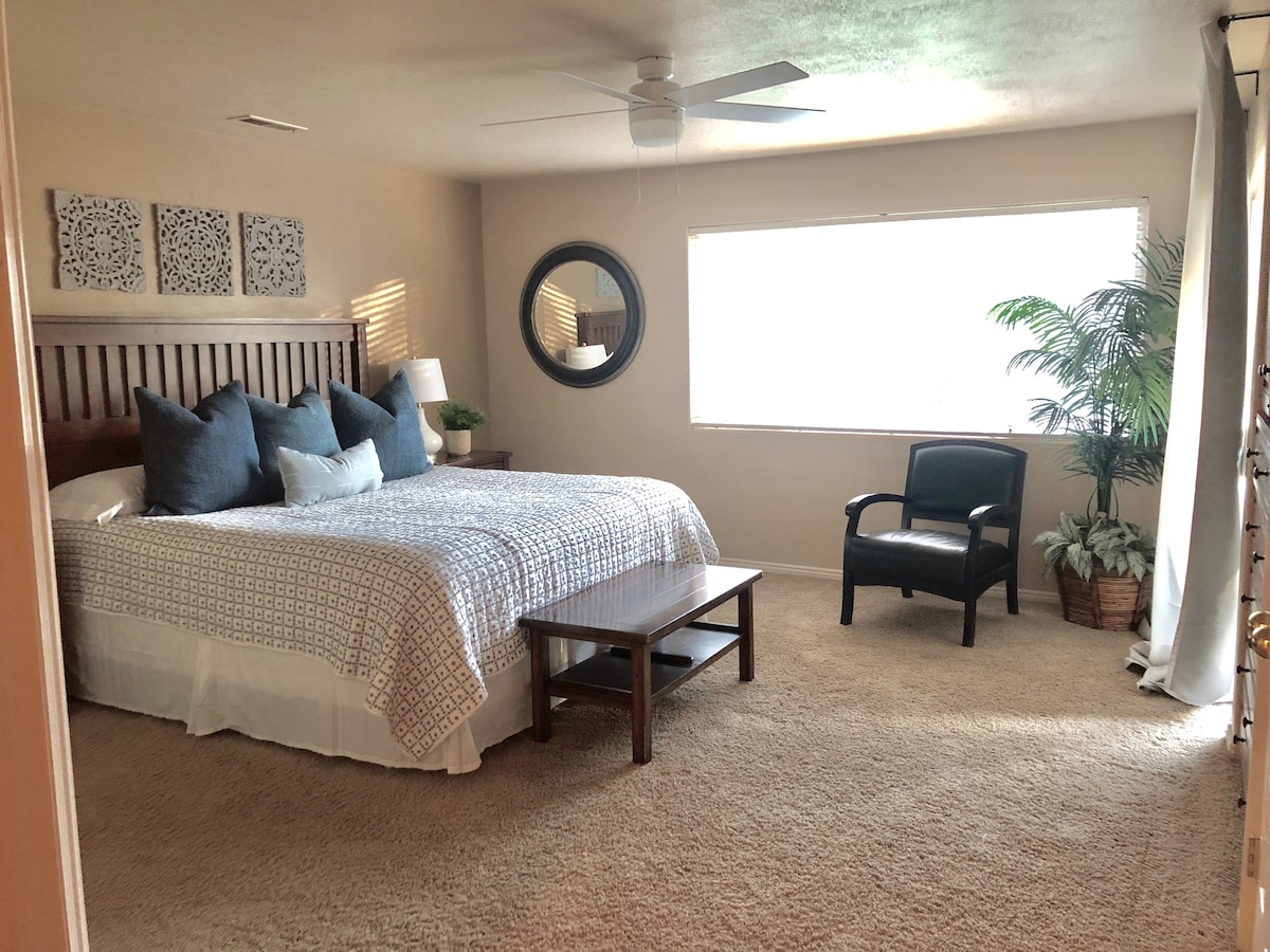 The spacious master bedroom features a king bed with a soft patterned blanket, accompanied by two large pillows and decorative throw pillows. A small coffee table is positioned in front of the bed. A comfortable chair sits beside a window, with natural light illuminating the room, complemented by a potted plant.