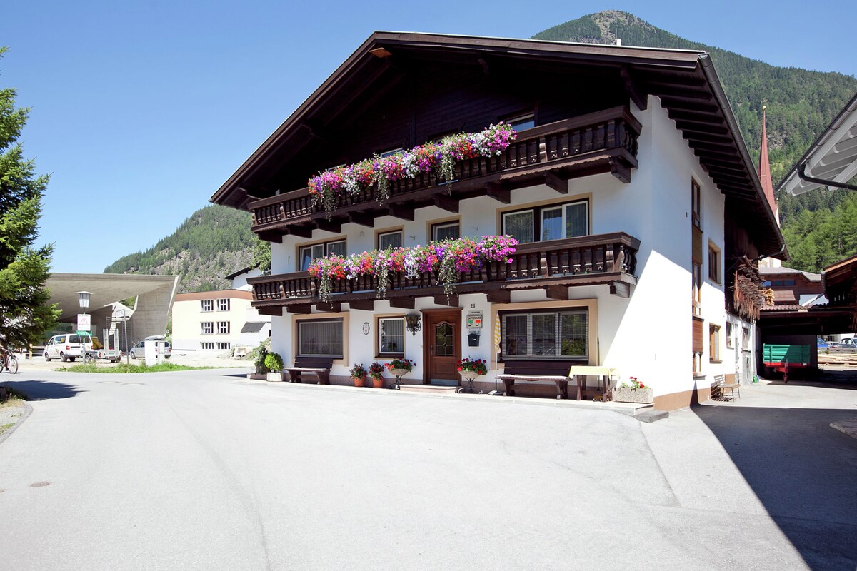 A two-story building features traditional alpine architecture with wooden balconies adorned with colorful flower boxes. The surrounding area includes a paved driveway and lush green mountains in the background, contributing to a serene setting. Ample parking space is visible in front of the structure.