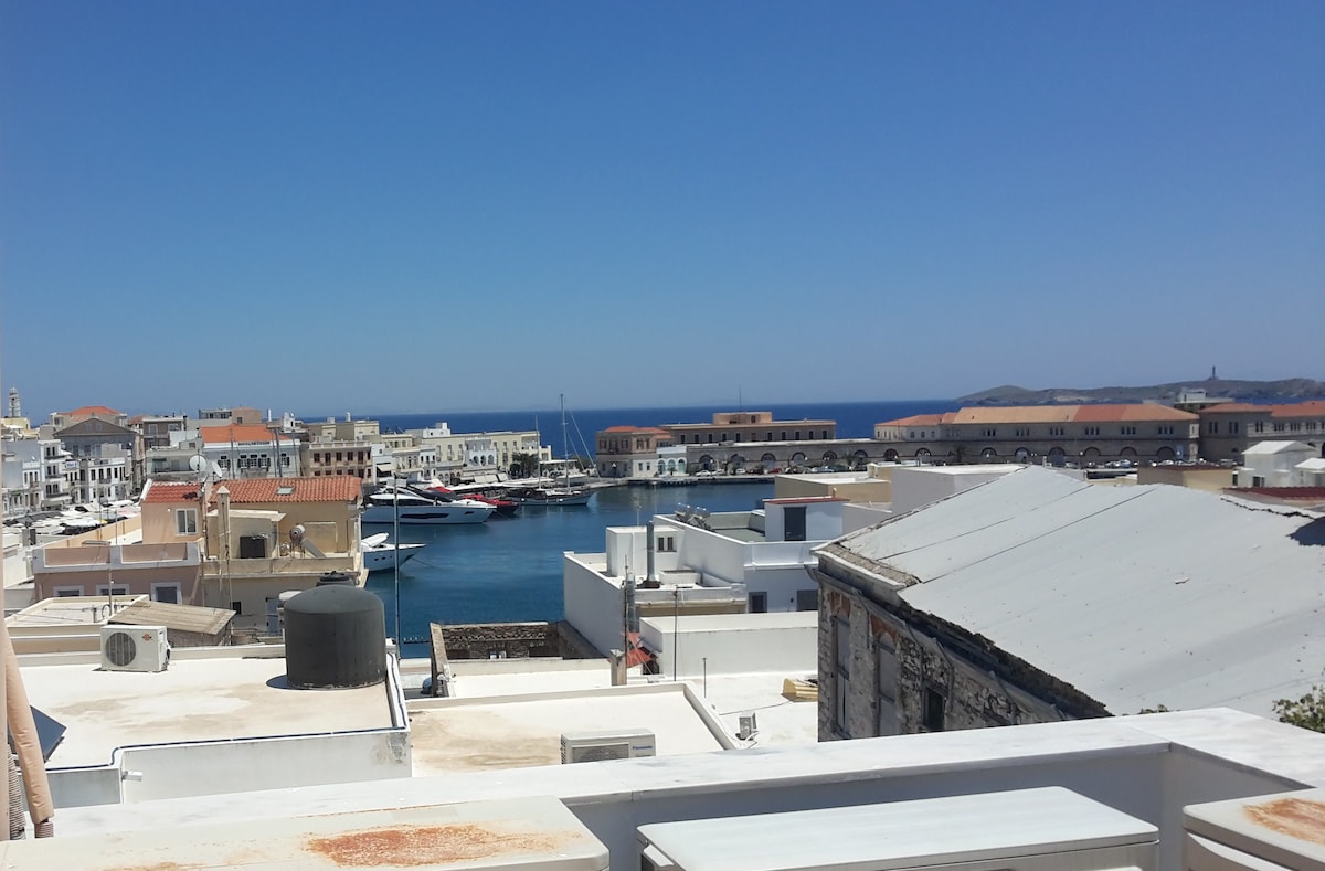 A view from the property showcases a picturesque harbor surrounded by various buildings. Boats are anchored in the calm waters, while the bright blue sea contrasts against the clear sky. The charming architecture of Ermoupolis can be seen, creating a vibrant coastal atmosphere.