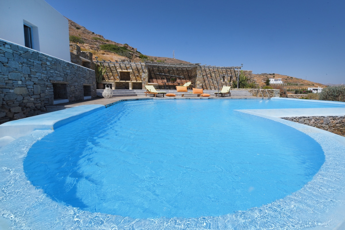 The inviting pool area features a large, curvilinear swimming pool with clear blue water. Surrounding loungers with cushions are positioned for relaxation. Stone walls provide a rustic backdrop, while the landscape of hills and structures in the distance creates a serene environment.