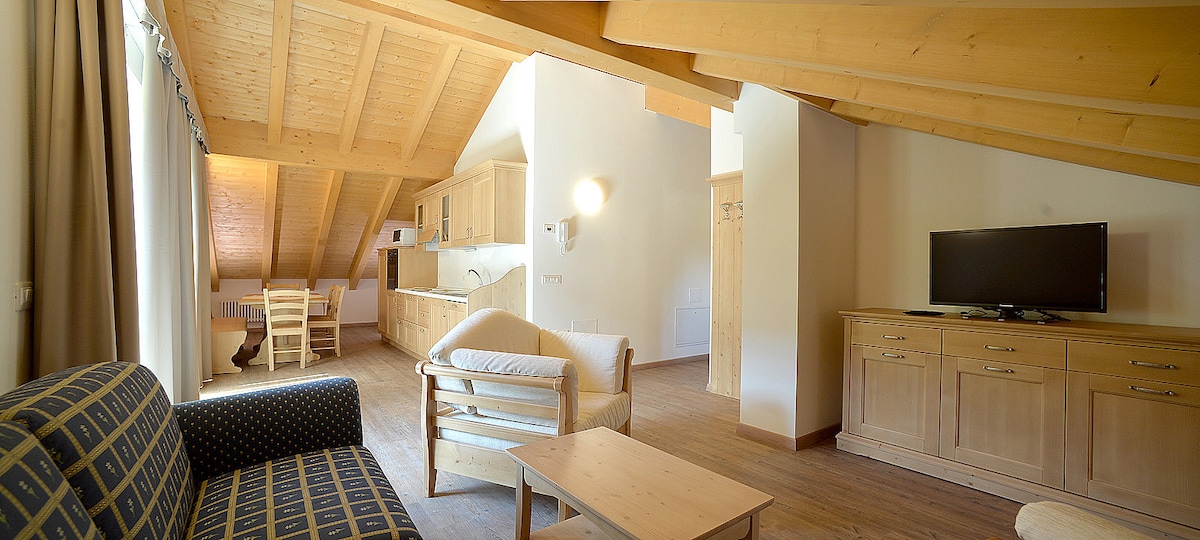 An open-concept living space showcases a light wood ceiling with exposed beams. A sofa and armchair are arranged beside a wooden coffee table, while a TV stands against a beige wall. The kitchen area features wood cabinetry, and natural light enters through large windows.