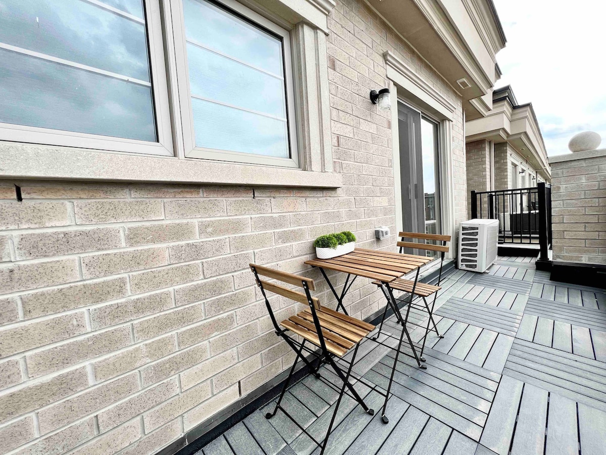 A small outdoor seating area is visible, featuring a wooden bistro table and two folding chairs. The space is framed by a brick wall and includes an air conditioning unit. Light-colored flooring adds a modern touch, while windows allow for natural light.