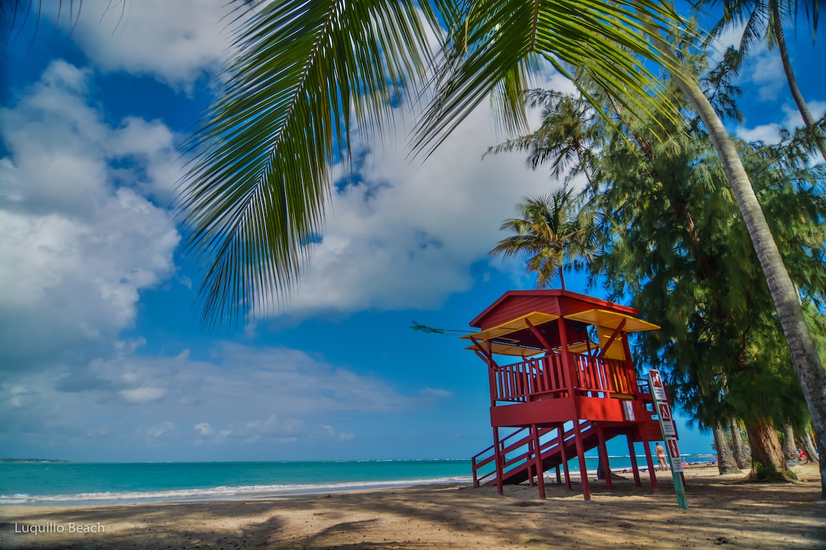 A bright red lifeguard tower stands along the sandy shore of Luquillo Beach, framed by palm trees swaying gently. The turquoise waters stretch into the horizon under a partly cloudy sky, creating a serene coastal atmosphere.