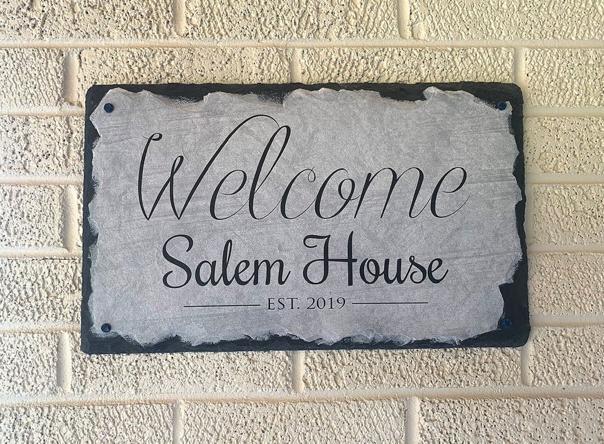 A welcome sign, crafted from stone, is mounted on a textured wall. The sign features the words 'Welcome Salem House' in elegant lettering, with 'EST. 2019' displayed below. The neutral tones of the stone complement the surrounding brick.