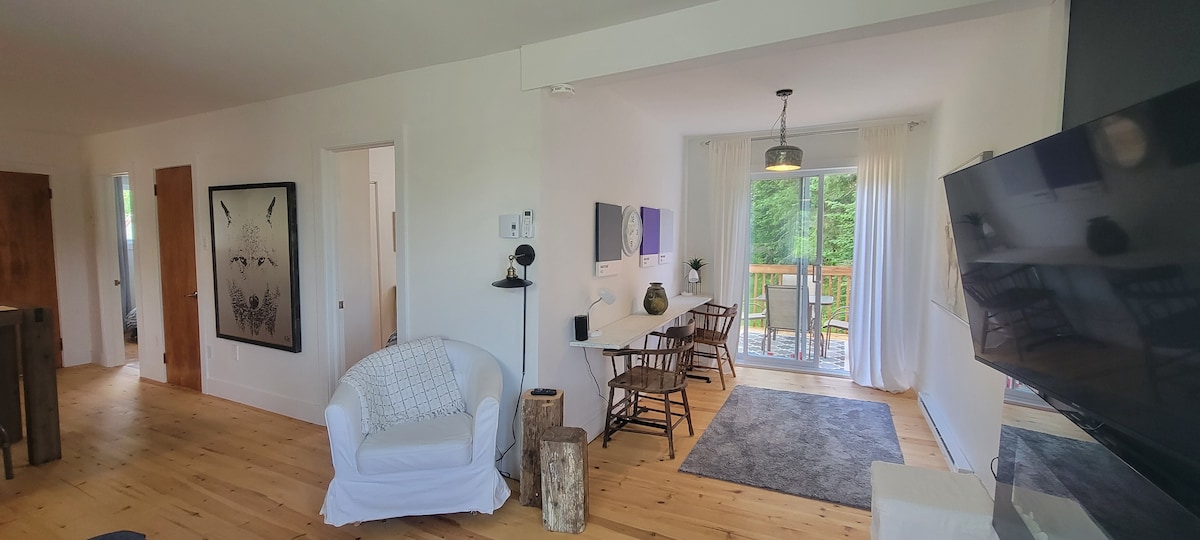 A welcoming living area features light wooden flooring and white walls. A cozy armchair is placed beside a television. A dining space contains a rectangular table with four chairs, complemented by natural light from large glass doors leading to a deck surrounded by greenery.