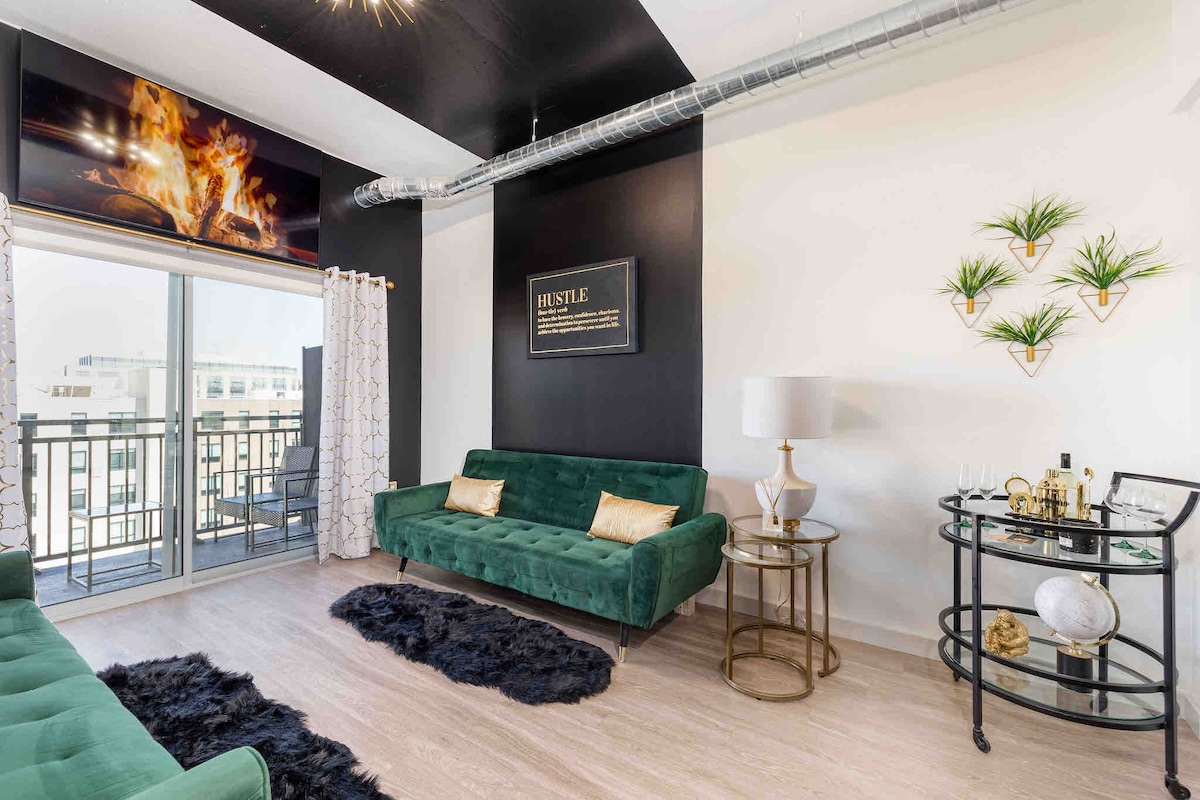 A stylish living area features a vibrant green couch paired with a matching loveseat, accented by gold cushions. A sleek, round coffee cart holds decorative items. Natural light fills the room through sliding glass doors leading to a balcony, with framed artwork on the dark wall.