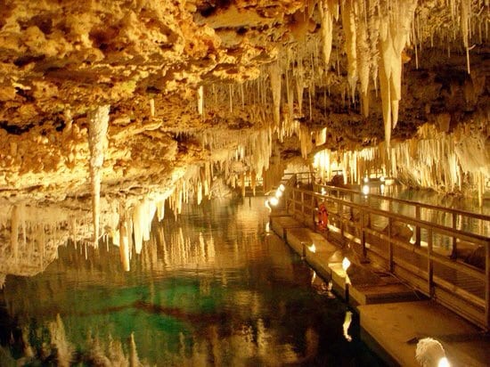 An underground cave display is characterized by hanging stalactites and a reflective pool of water. Soft lighting illuminates the area, highlighting the natural formations and creating an enchanting atmosphere. A wooden walkway is visible, allowing for easy exploration of this unique environment.