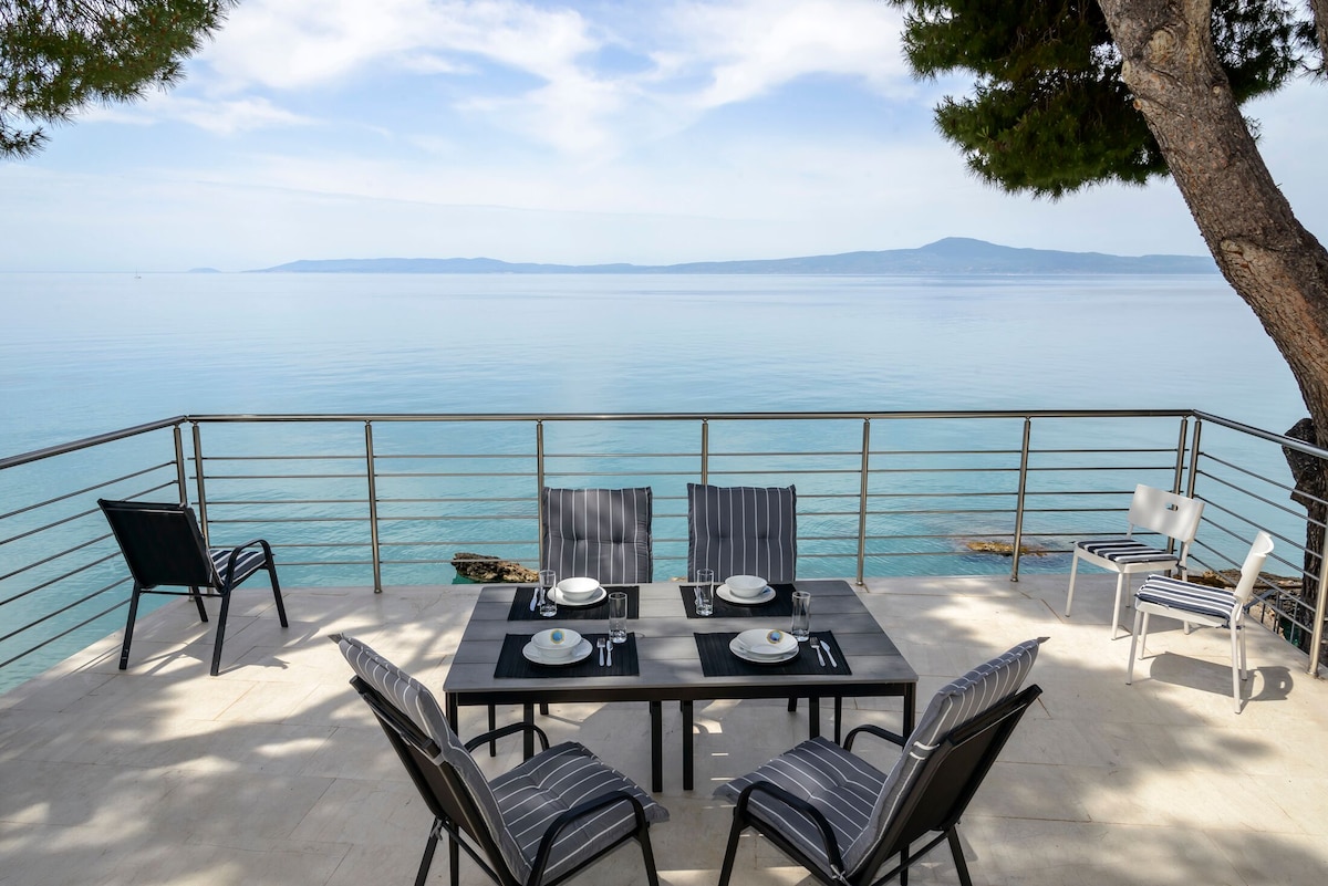 An outdoor dining area is situated on a spacious terrace, featuring a sleek table set with plates and cutlery. Surrounding chairs provide ample seating, while the serene sea view stretches to the horizon. A tree offers shade, enhancing the inviting atmosphere.