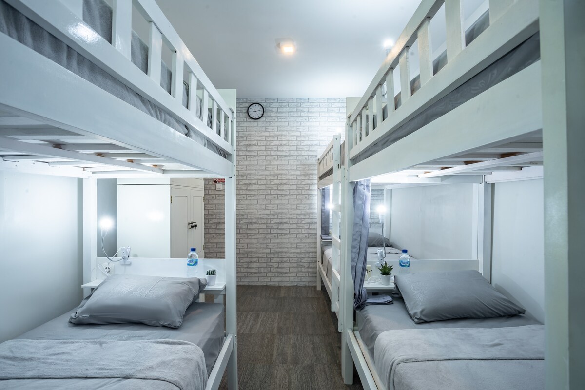 A shared bedroom is equipped with two sets of bunk beds, each featuring individual reading lights and small shelves. The walls are painted white, complemented by a brick accent wall. Neutral-colored bedding is neatly arranged on each bed, providing a calming atmosphere.