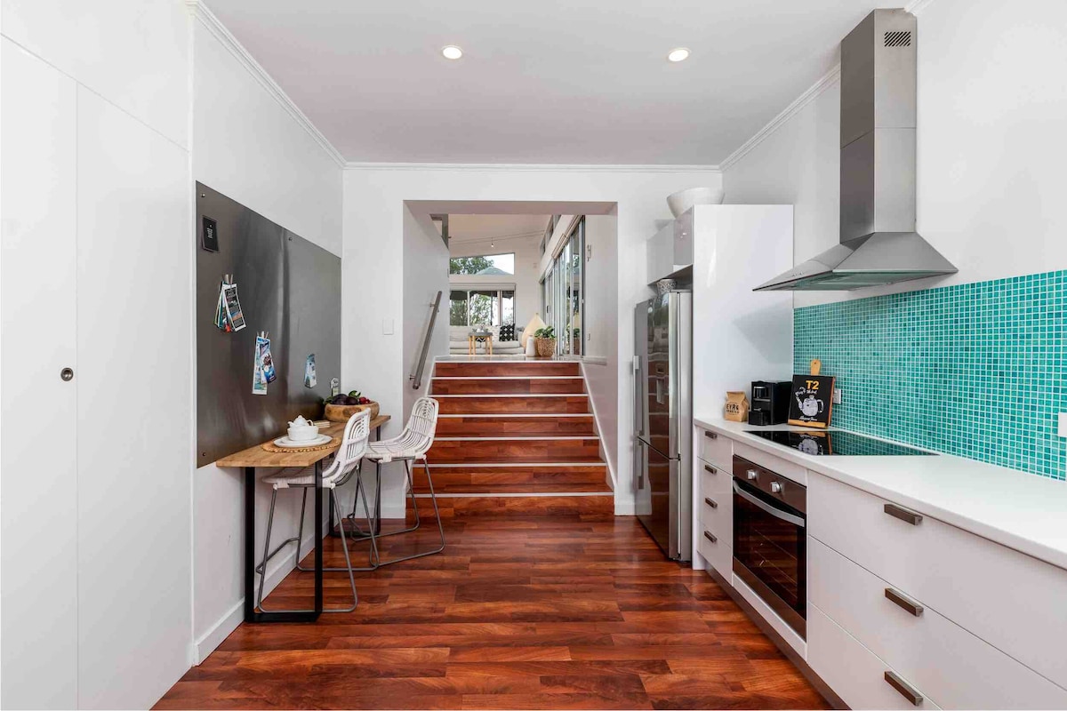 A spacious kitchen area features sleek cabinets, white countertops, and modern appliances. A vibrant turquoise tile backsplash adds a pop of color. A small dining table with white chairs is positioned near the entrance, leading to a staircase bathed in natural light.
