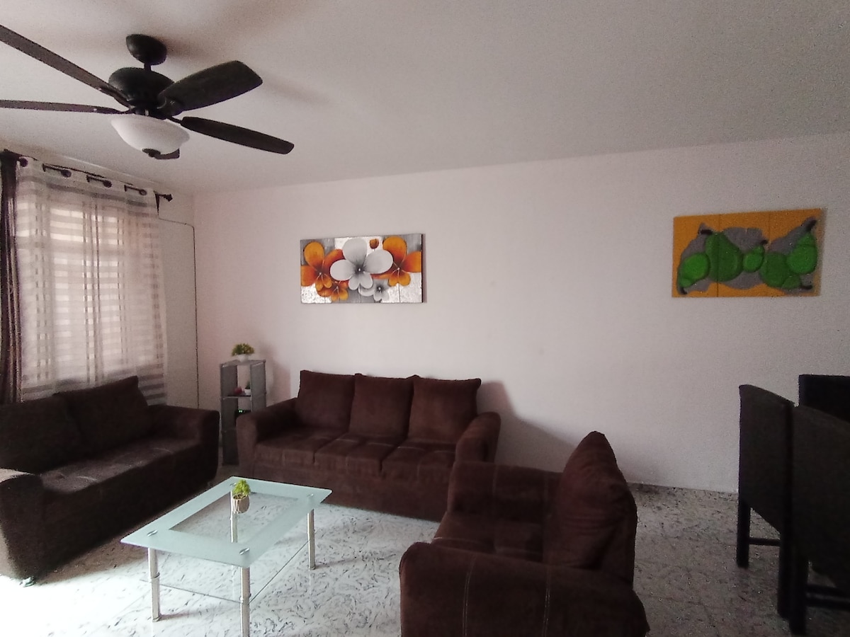 The living area features a comfortable seating arrangement with two dark brown sofas and a glass coffee table at the center. Complementary wall art pieces adorn the walls, and natural light filters through partially sheer curtains.