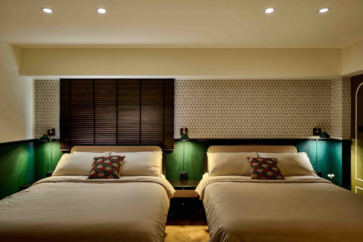 The room features two spacious beds dressed in neutral linens, complemented by decorative pillows. A decorative wall with geometric patterns and wooden blinds enhances the aesthetic, while soft ceiling lights provide a warm ambience.