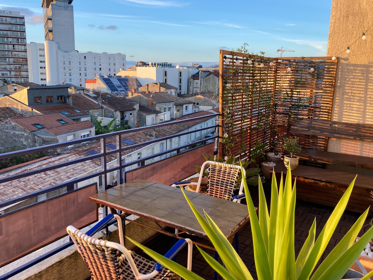 A spacious terrace is available featuring a table and chairs, surrounded by various plants. A view of Bordeaux's rooftops and distant buildings is visible, with natural light illuminating the space. The area is enclosed by wooden panels, creating an inviting outdoor setting.