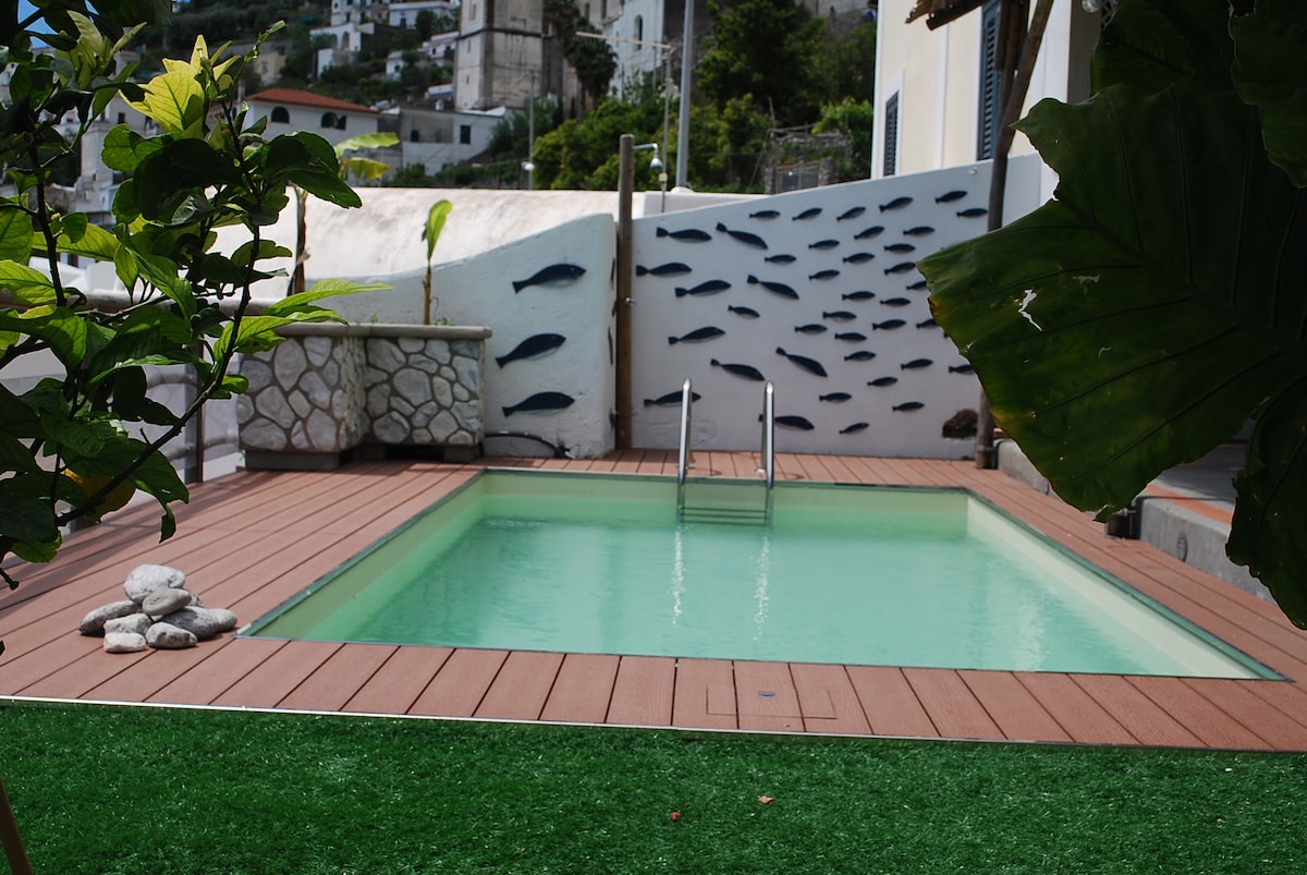 A private swimming pool is framed by wooden decking and greenery. The clear water invites relaxation, while a decorative wall with fish motifs adds a unique touch. The setting is complemented by a patch of artificial grass and stone accents, creating a serene outdoor space.