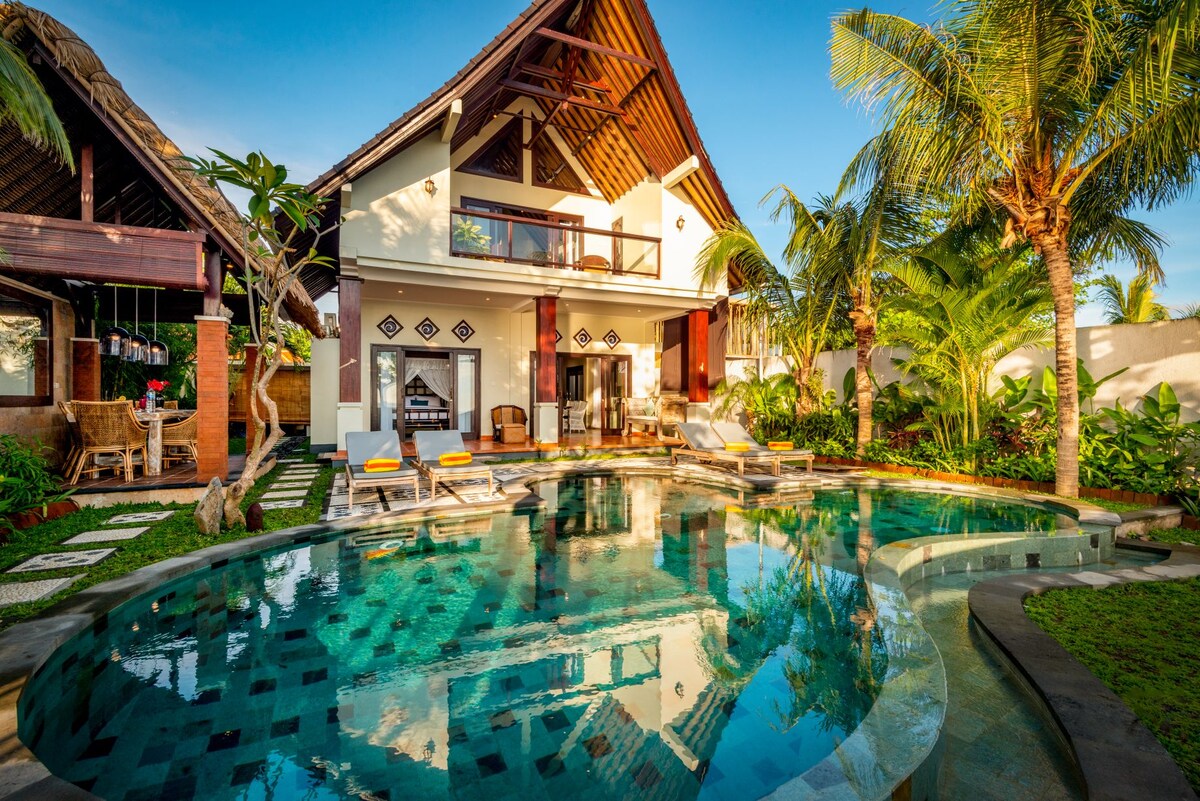 A private villa is surrounded by lush greenery, featuring a curved infinity pool that reflects the clear blue sky. Sun loungers are positioned along the pool deck, offering spacious areas for relaxation. The two-story villa boasts high ceilings and large glass doors, enhancing the sense of openness.