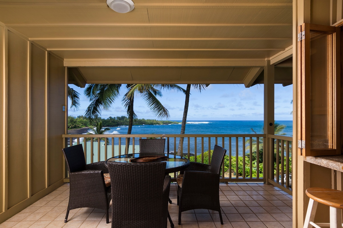 A covered lanai features a round dining table surrounded by four wicker chairs. Lush greenery and palm trees frame a panoramic view of the ocean, where gentle waves meet the shore under a partly cloudy sky.