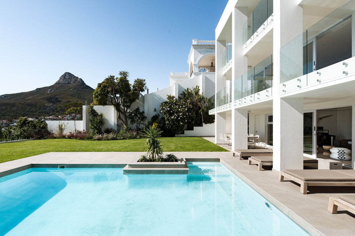 A spacious pool area is framed by manicured lawns and lush greenery, with a clear blue pool reflecting the sky. Modern sun loungers line the poolside, while the villa's sleek architecture and mountain views create a serene atmosphere.