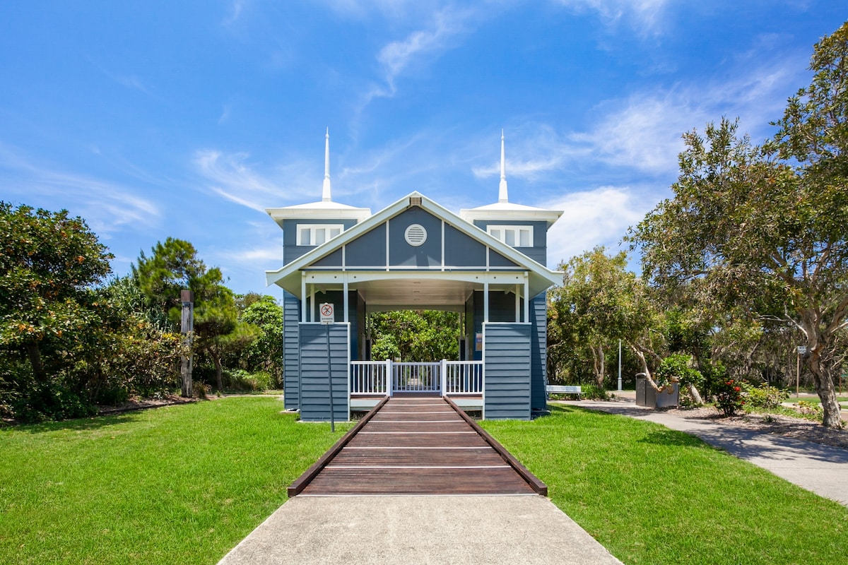 A charming cottage exterior features a wide wooden walkway leading to a covered entrance. The structure displays a blue façade, accentuated by white detailing and twin gables. Lush greenery surrounds the home, contributing to a serene atmosphere and inviting approach.