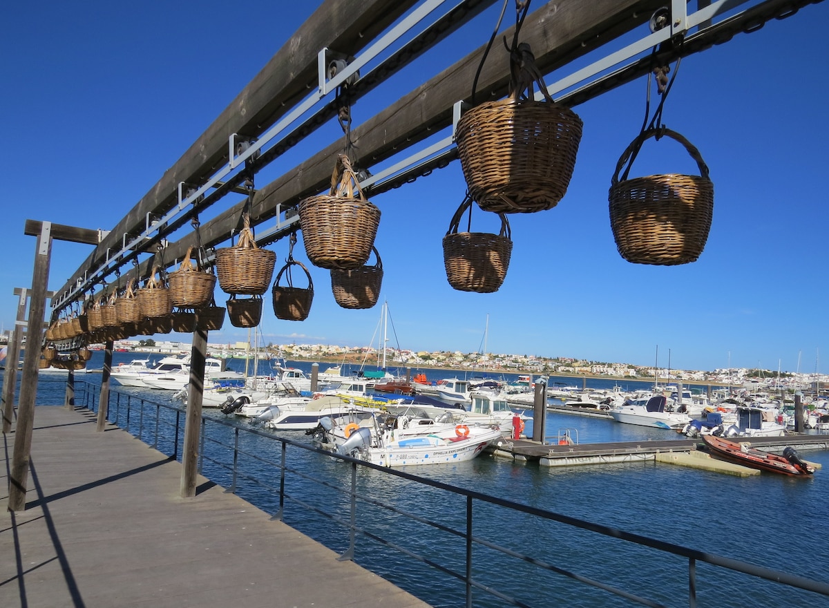 A row of woven baskets hangs suspended on a wooden beam above a marina. Below, various boats are docked in the calm water, with the coastline visible in the distance under a clear blue sky.