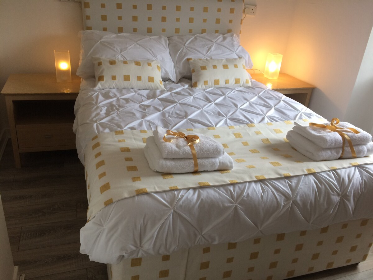 A well-appointed sleeping area features a spacious bed adorned with a patterned yellow and white spread. Fluffy towels are neatly arranged on the bed, and soft lighting is provided by two bedside lamps, creating a calm and relaxing environment.