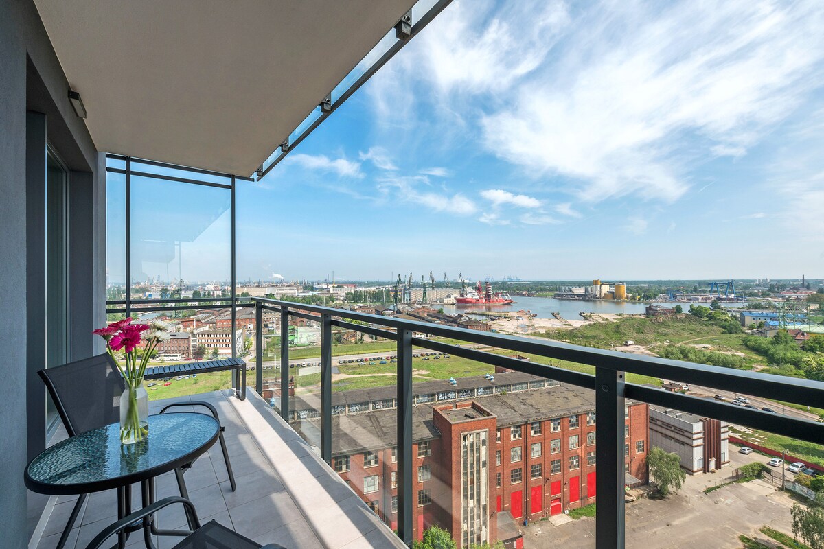 A balcony features a glass railing, providing views of the Old Town and the Shipyard. A round table with a vase of flowers is placed on the tiled floor, set against a backdrop of the vibrant skyline and waterfront.