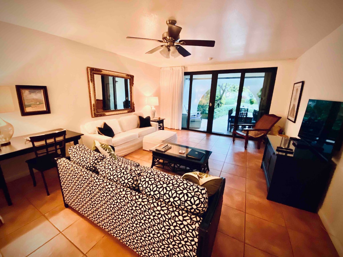 A living area features a contrasting black and white patterned couch and a light-colored sofa, complemented by a wooden coffee table. Large glass doors provide views to the outside seating area. Artwork adorns the walls, and a ceiling fan adds to the comfort.