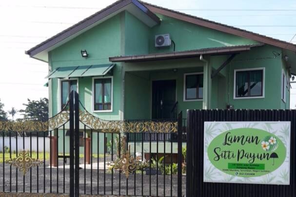 A green two-story house is depicted, featuring a welcoming entrance with steps and a shaded doorway. The property is enclosed by a decorative black gate, while a sign displaying the name 'Laman Sitti Payun' is prominently placed at the front, highlighting the cozy ambiance of the location.