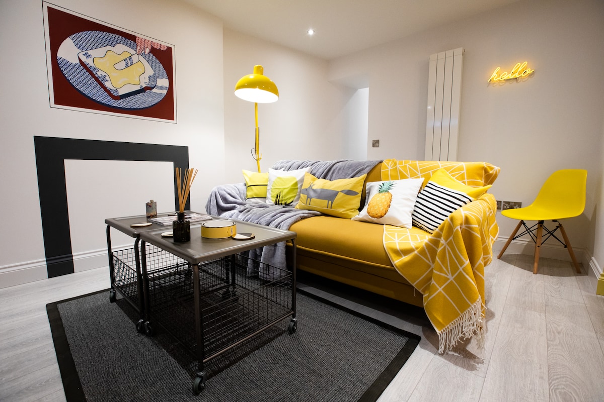 The inviting living area features a yellow sofa adorned with colorful cushions and a patterned throw. A modern coffee table occupies the center, accompanied by decorative items. A neon sign and framed artwork enhance the aesthetic, while soft lighting creates a cozy ambiance.