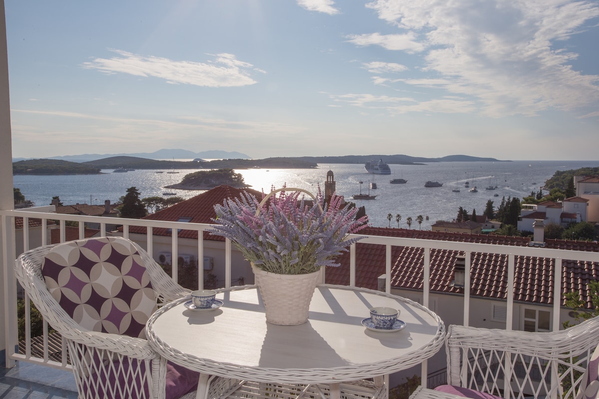 A balcony table is set with two cups and a small vase of lavender. The view features shimmering waters with boats and islands in the distance, highlighting the coastal landscape under a clear sky.