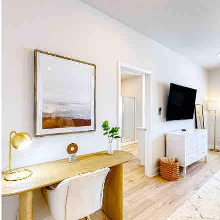 A well-lit workspace features a wooden desk adorned with a lamp and decorative plant. A stylish chair sits in front of the desk, with artwork on the wall and a television mounted nearby. A light area rug adds texture to the warm wood floor.