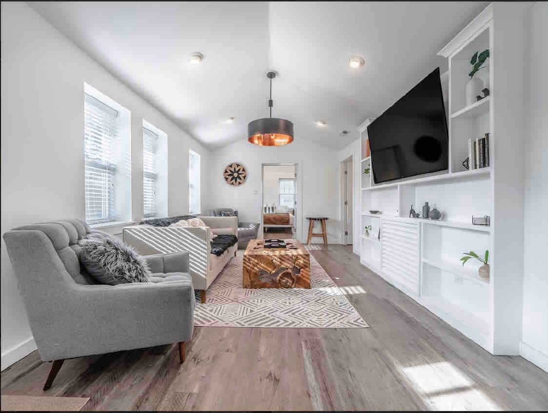 A modern living area features large windows allowing natural light to brighten the space. A stylish gray sofa complements a striped chair, while a rustic coffee table sits on a patterned rug. A mounted television is visible above a minimalistic entertainment unit.