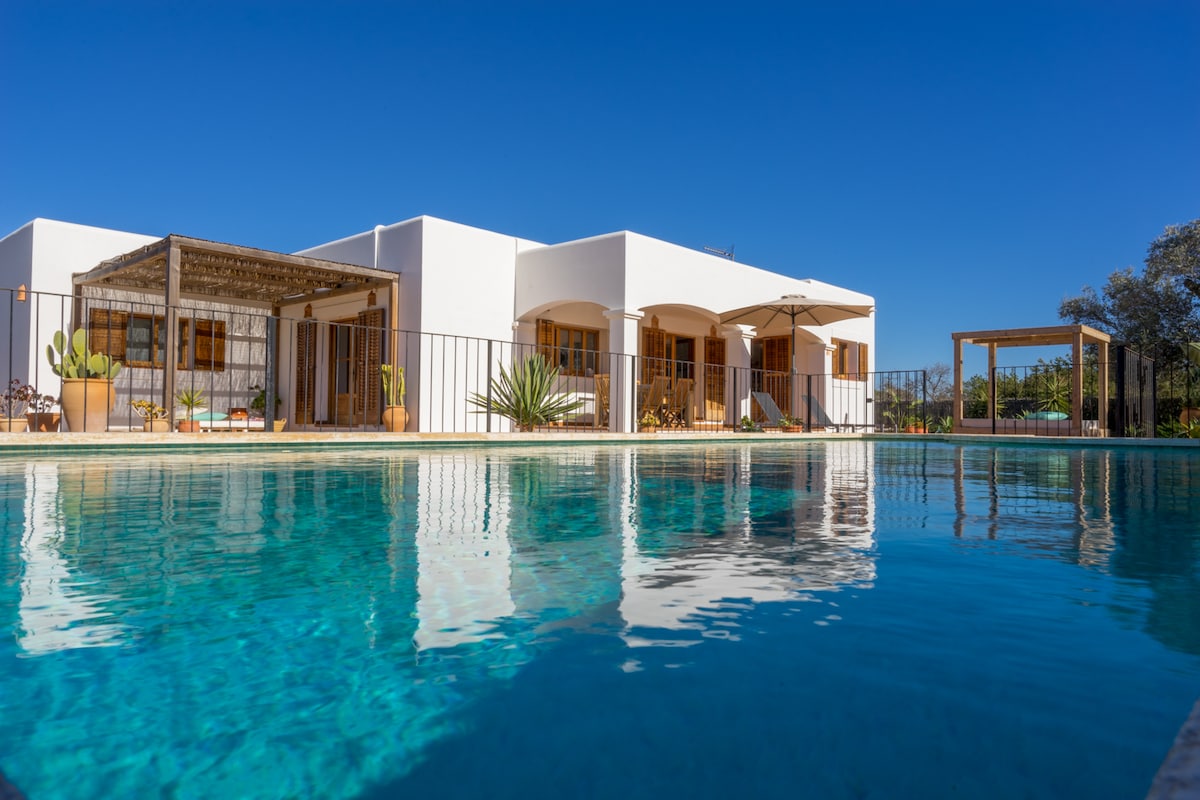 The villa features a white exterior with arched entrances, set against a clear blue sky. The tranquil pool reflects the surrounding landscape and offers a serene environment. Large windows and shaded areas provide both light and comfort for outdoor relaxation.