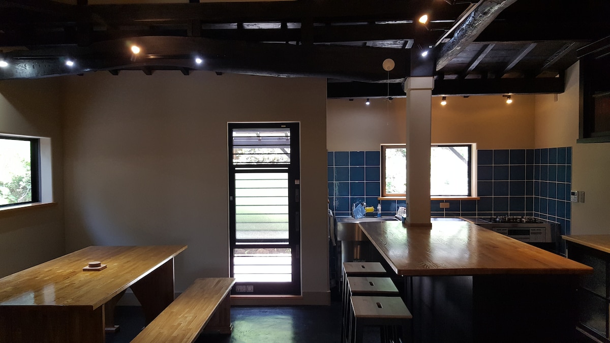 An airy kitchen space features a large wooden table with bench seating, complemented by bar stools positioned at a wooden countertop. Natural light enters through two windows and a door, creating a bright atmosphere. A blue tiled wall adds a pop of color, enhancing the area.