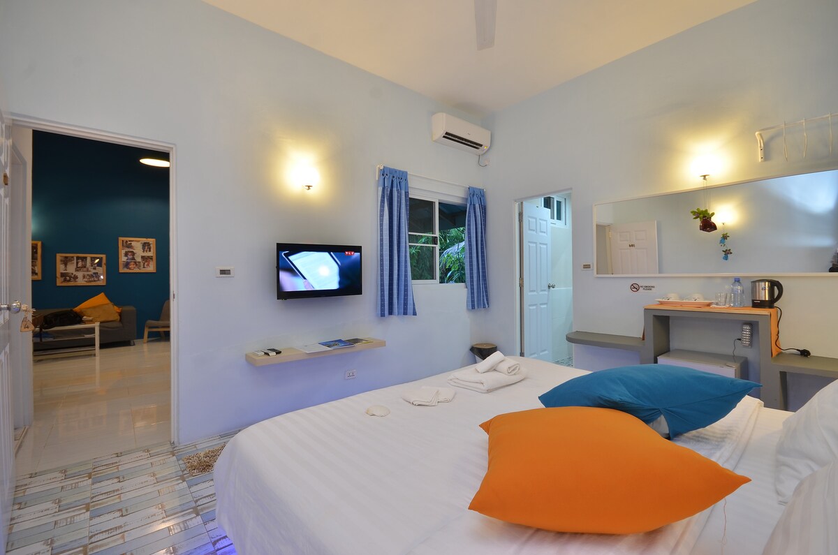 A spacious guest room features a large bed adorned with colorful pillows. A wall-mounted television is visible, alongside a window framed by light blue curtains. A small kitchenette area is seen in the background, and a door leads to another room.