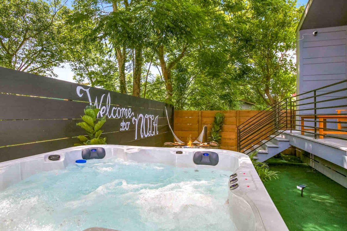 A hot tub is situated amidst a lush green outdoor space, surrounded by a privacy fence that enhances the tranquil setting. Natural elements, including plants, complement the inviting atmosphere. A welcoming sign is displayed on the fence, adding a personal touch to the area.